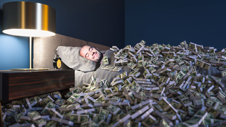 © Getty Images/iStockphoto Smiling man sleeping in a bed covered with dollars money. wealth concept.