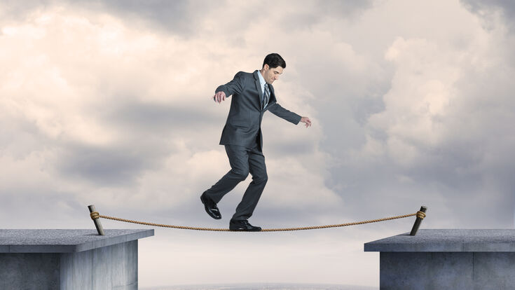 © Getty Images A businessman balancing on a tightrope high above a big city. He looks down at the rope before his next step as he nears the midpoint of the tightrope. Metal spikes are anchored into the concrete to help support the tightrope. The New York City skyline can be seen in the distance as a fog persists over the buildings.