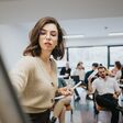 A young woman leads a presentation at a business workshop, confidently engaging with a group of professionals seated in a stylish office setting.