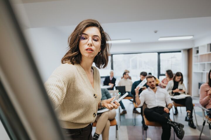 A young woman leads a presentation at a business workshop, confidently engaging with a group of professionals seated in a stylish office setting.