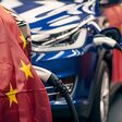 A line of electric cars are being charged at an event, with a Chinese flag in the foreground. The automotive design, wheels, and electric blue colors stand out