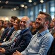 Business people happily listen to startup owner at trade show. Laughing, engaged. Event in large hall. Atmosphere lively enthusiastic. Business people seem interested in presentation. Many people in