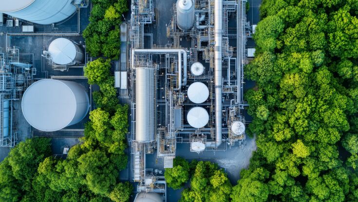 © VERTEX SPACE - stock.adobe.com Aerial view of an industrial facility surrounded by lush green trees, showcasing blend of nature and industry. image captures intricate network of pipes and storage tanks.