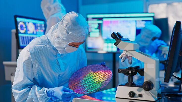 asian male technician in sterile coverall holds wafer that reflects many different colors with gloves and check it at semiconductor manufacturing plant