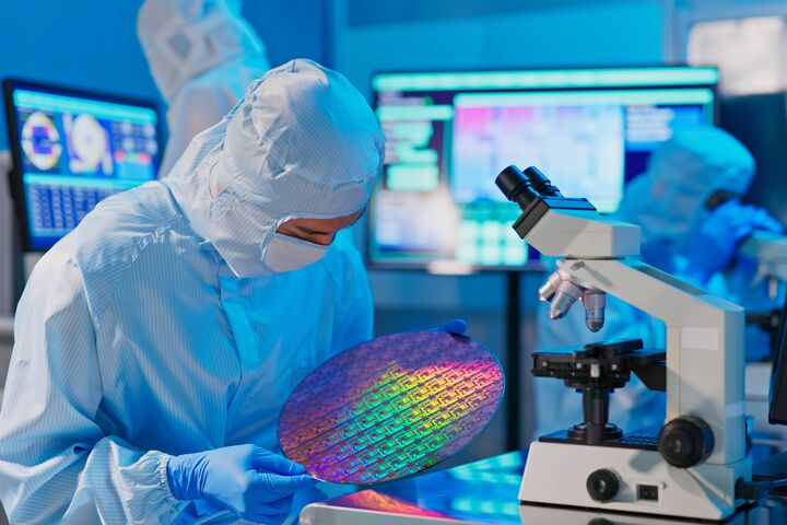 asian male technician in sterile coverall holds wafer that reflects many different colors with gloves and check it at semiconductor manufacturing plant