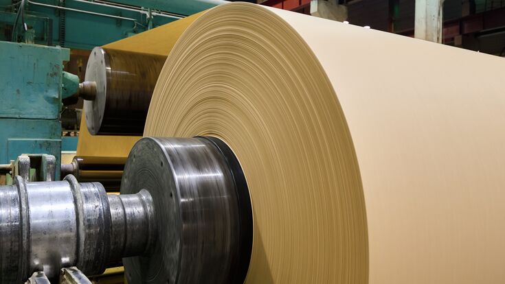 A massive roll of smooth brown paper is being processed in a busy paper manufacturing plant, showcasing the machinery in operation.