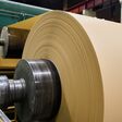 A massive roll of smooth brown paper is being processed in a busy paper manufacturing plant, showcasing the machinery in operation.