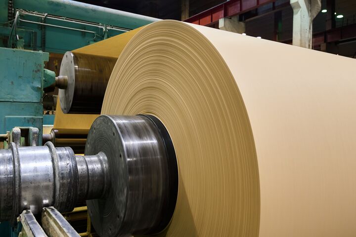 A massive roll of smooth brown paper is being processed in a busy paper manufacturing plant, showcasing the machinery in operation.