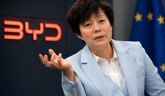 BYD-Managerin vor BYD-Logo und EU-Flagge bei einem Gespräch über die Europa-Strategie des chinesischen Elektroautobauers.