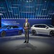 BMW präsentiert die Neue Klasse auf einer Bühne mit zwei Elektroautos, darunter ein weißer BMW iX3, vor großem Schriftzug „The Neue Klasse. Start into a new era“.