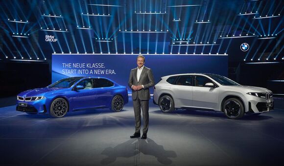BMW präsentiert die Neue Klasse auf einer Bühne mit zwei Elektroautos, darunter ein weißer BMW iX3, vor großem Schriftzug „The Neue Klasse. Start into a new era“.