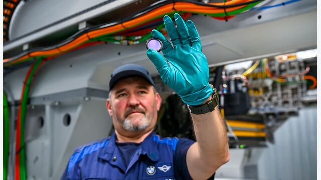 BMW-Mitarbeiter hält eine Batteriezelle in einer technischen Fertigungsumgebung mit Hochvoltkabeln.