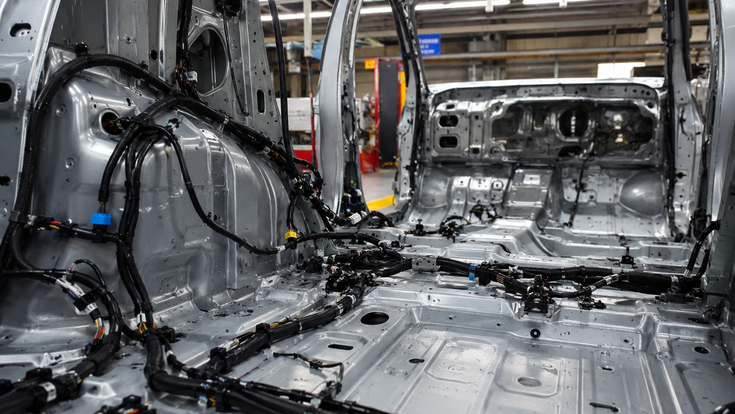 Innenansicht einer Rohkarosse in einer Autofabrik. Mehrere schwarze Kabelbäume und Bordnetzleitungen sind entlang des Fahrzeugbodens und der Seitenwände verlegt.