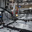 Innenansicht einer Rohkarosse in einer Autofabrik. Mehrere schwarze Kabelbäume und Bordnetzleitungen sind entlang des Fahrzeugbodens und der Seitenwände verlegt.