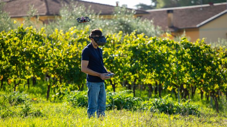 Man wearing FPV goggles and holding a remote control flying a small drone over a lush green vineyard, exploring new perspectives with advanced technology in nature