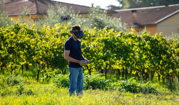 Man wearing FPV goggles and holding a remote control flying a small drone over a lush green vineyard, exploring new perspectives with advanced technology in nature