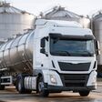 A white tanker truck waits at an industrial facility beside storage silos as workers inspect equipment.